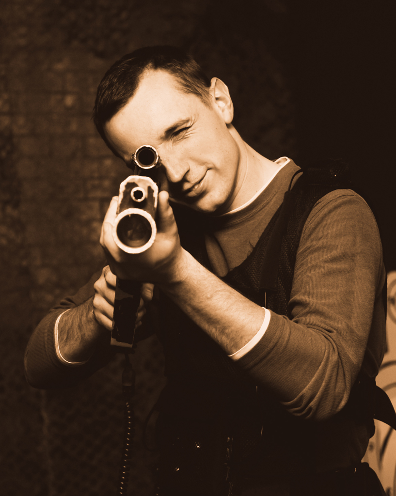 Mężczyzna w kamizelce celuje z pistoletu laserowego, przymrużając oko, w pomieszczeniu o ciemnym, ceglanym tle.