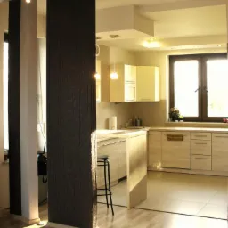 Jasna kuchnia z barkiem i wysokim stołkiem, oddzielona od salonu dekoracyjną kolumną w odcieniach szarości. Parkiet na podłodze, okno z kwiatami w tle.