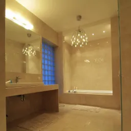 Jasna łazienka z beżowymi płytkami, nowoczesną umywalką i wanną. Niebieskie szklane luksfery w oknie wpuszczają światło. Designerska lampa sufitowa.