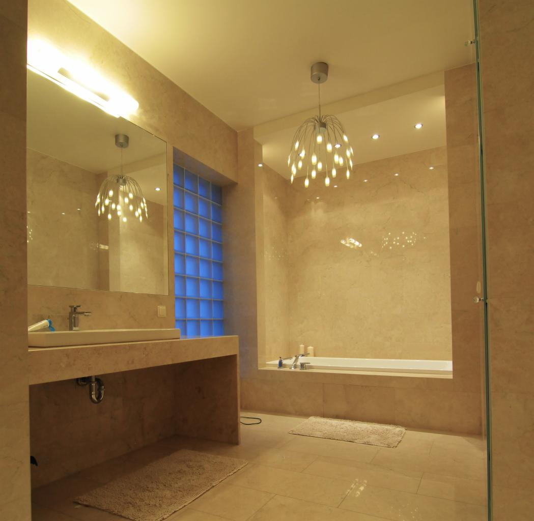 Jasna łazienka z beżowymi płytkami, nowoczesną umywalką i wanną. Niebieskie szklane luksfery w oknie wpuszczają światło. Designerska lampa sufitowa.