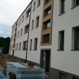 Odnowiona elewacja budynku wielorodzinnego z fragmentami wykończenia cegłą klinkierową, widoczne materiały budowlane zabezpieczone folią w pasy.