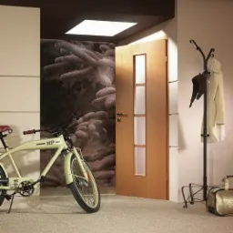 Otwarta drewniana drzwi wewnętrzne z trzema szybami, widok na ścianę z fototapetą przedstawiającą gałązki drzew iglastych, obok beżowy rower i wieszak na ubrania w nowoczesnym przedpokoju.