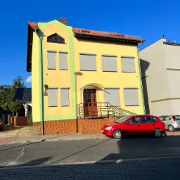 Żółty dom z zielonymi akcentami i szarymi roletami zewnętrznymi, czerwony samochód zaparkowany przed budynkiem.