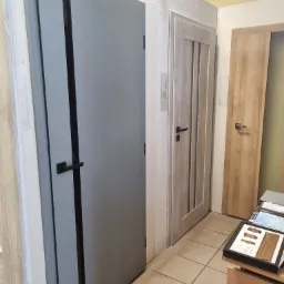 Wystawa trzech modeli drzwi wewnętrznych: szare, z przeszkleniami i drewnopodobne, z czarnymi i srebrnymi klamkami, na tle płytek podłogowych.