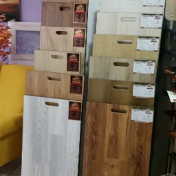 Ekspozycja próbek paneli podłogowych różnych kolorów i wzorów, marki Alpina Floor, z uchwytami i informacją o 25-letniej gwarancji.