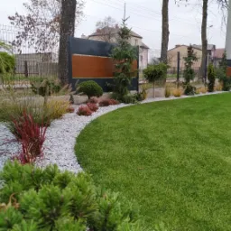 Nowoczesny ogród z równo przystrzyżonym trawnikiem, obrzeżem z białego grysu, ozdobnymi trawami i kulistą lampą ogrodową, na tle metalowo-drewnianego ogrodzenia i drzew.