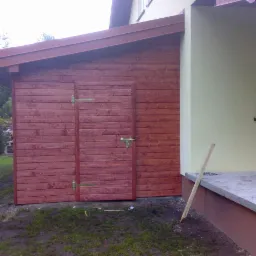 Drewniana dobudówka z dwuskrzydłowymi drzwiami, pomalowana na czerwono-brązowy kolor, przylegająca do ściany budynku z małym oknem. Widoczna młoda sosna w tle.