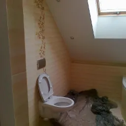 Łazienka na poddaszu z oknem dachowym, wykończona beżowymi płytkami z motywem roślinnym, biała podwieszana toaleta z otwartą klapą, podłoga zabezpieczona folią malarską.