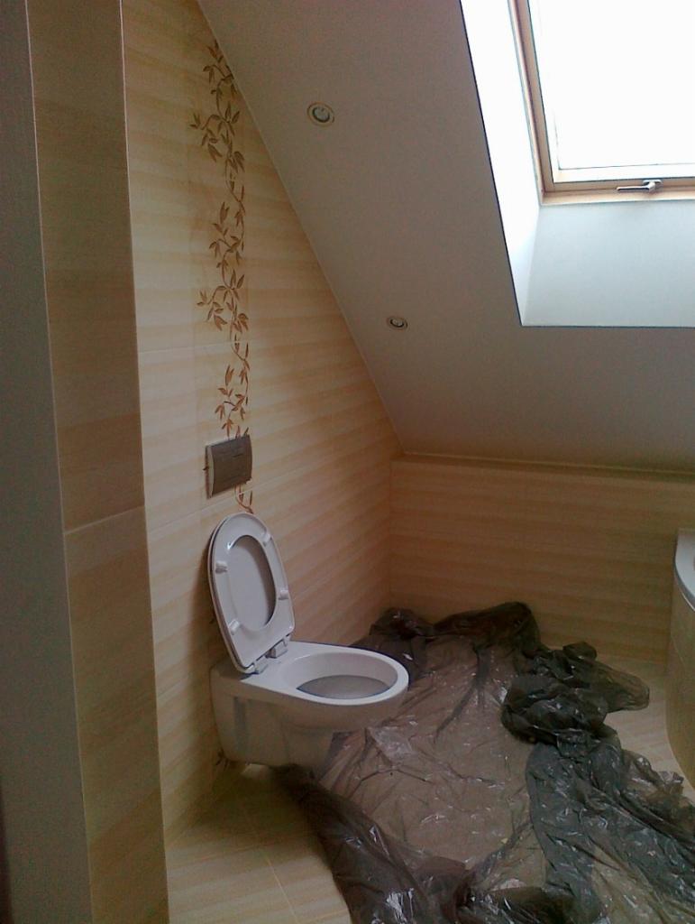 Łazienka na poddaszu z oknem dachowym, wykończona beżowymi płytkami z motywem roślinnym, biała podwieszana toaleta z otwartą klapą, podłoga zabezpieczona folią malarską.