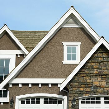 Fragment elewacji domu z nałożonym tynkiem akrylowym w kolorze brązowym, z widoczną fakturą 'baranka', połączony z elementami z kamienia naturalnego i białymi ramami okiennymi.