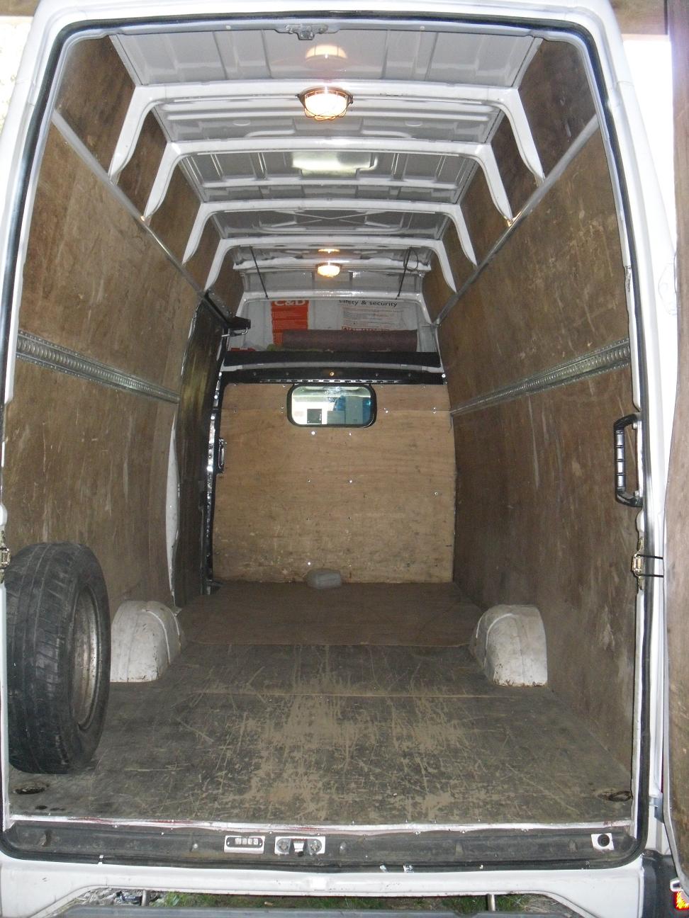 Wnętrze furgonetki wyłożone sklejką, z zamontowanymi szynami mocującymi i zapasowym kołem po lewej stronie, oświetlone lampami sufitowymi.