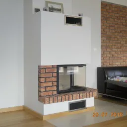 Nowoczesny kominek z obudową z cegły i białym wykończeniem, wkomponowany w ścianę z cegły dekoracyjnej, obok skórzanej sofy.