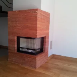 Nowoczesny kominek narożny z obudową z czerwonego trawertynu, widok na przeszklone palenisko, drewniana podłoga, minimalistyczne wnętrze.