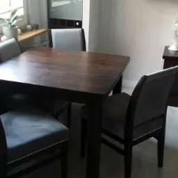 Ciemny, drewniany stół z szarymi krzesłami w jadalni, widok na okno z dekoracyjnymi kulami świetlnymi i komodę z lampką.