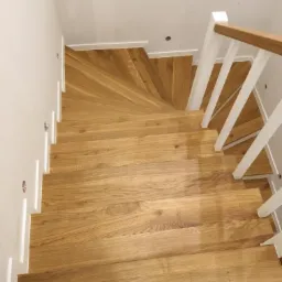 Widok z góry na drewniane schody z jasnego drewna z białymi balustradami i listwami przypodłogowymi, z widocznym zakrętem na półpiętrze.