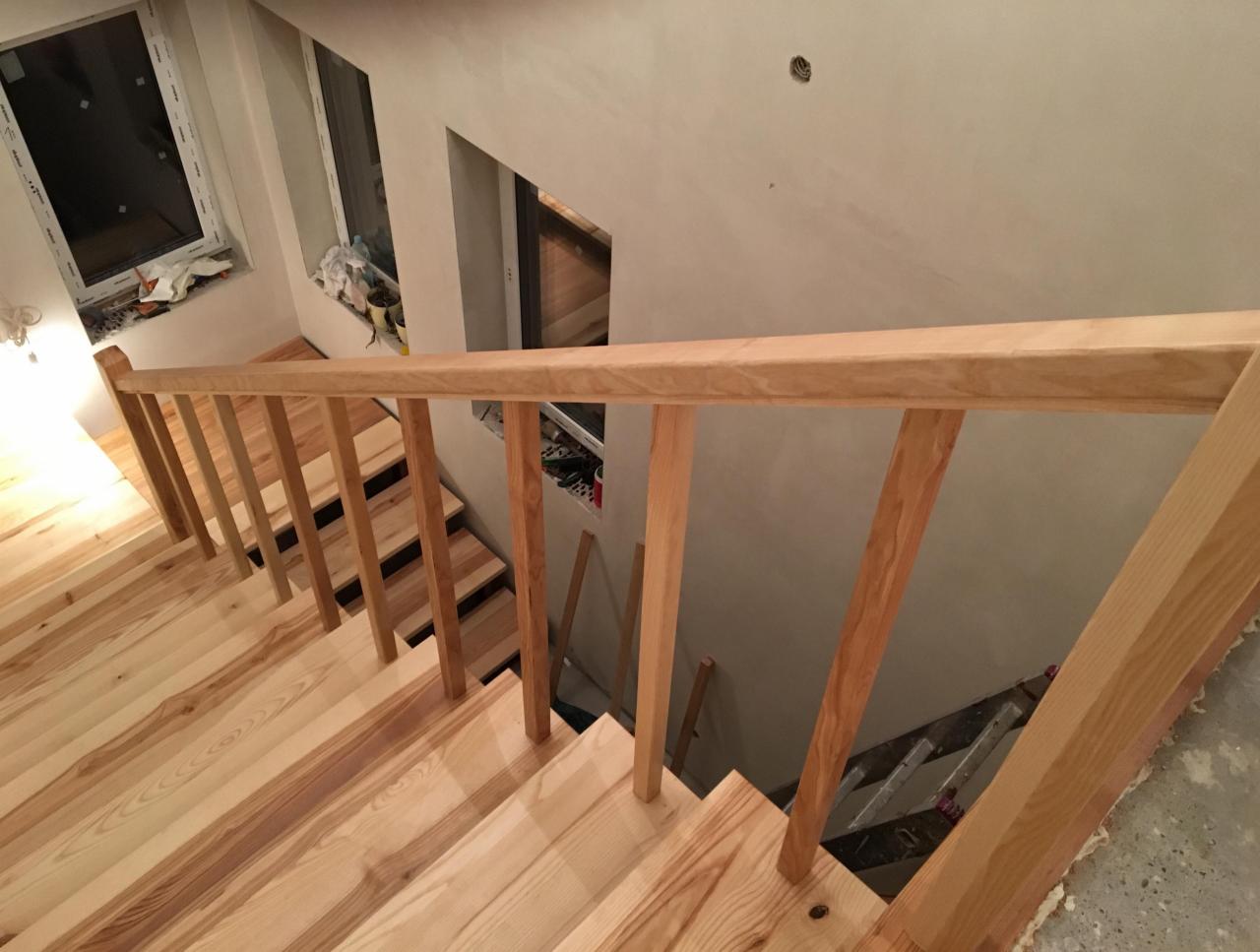 Jasne drewniane schody z balustradą w trakcie montażu, widoczne niedokończone wykończenie ścian i okien