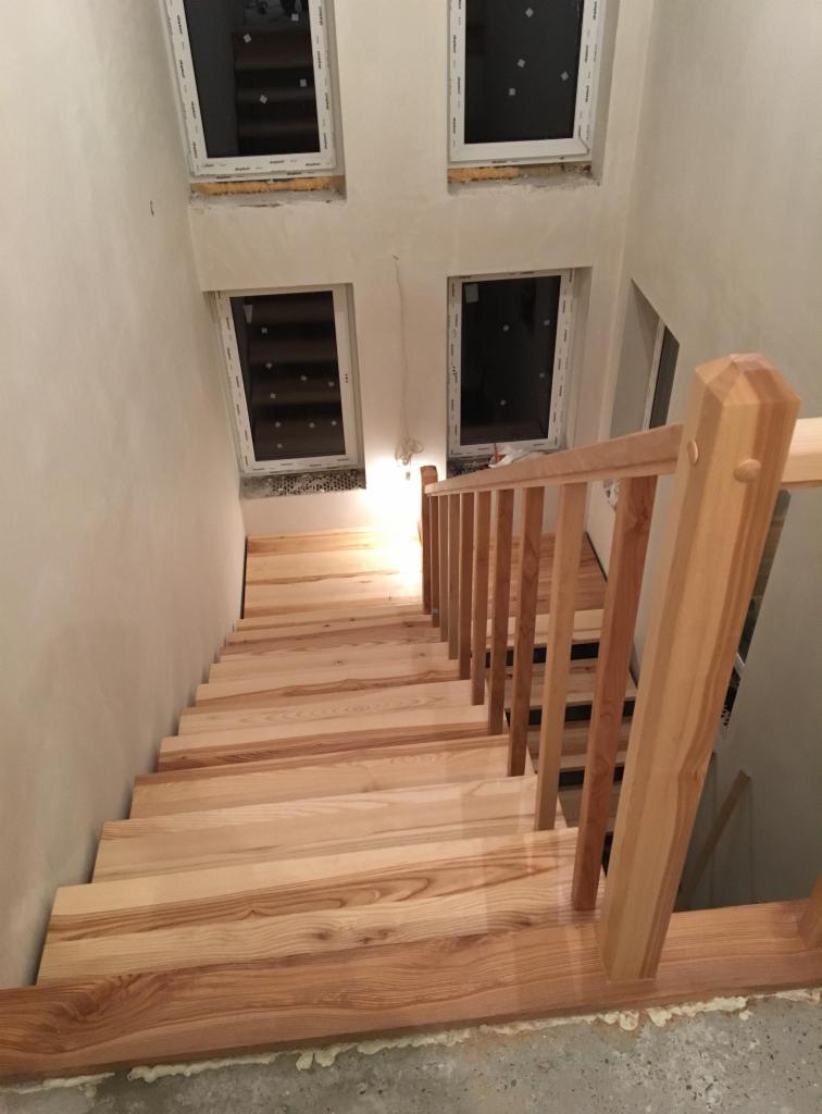 Jasne, drewniane schody z poręczą, prowadzące w dół, z widocznymi czterema oknami dachowymi w białych ramach i surowymi ścianami.