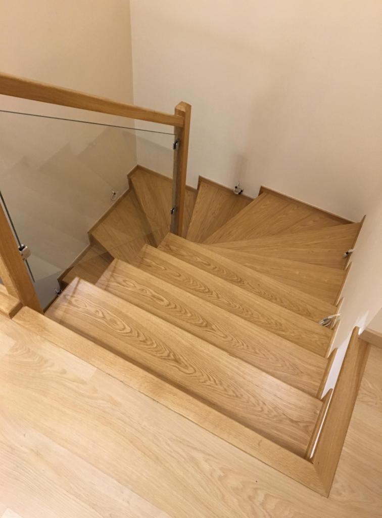 Drewniane schody spiralne z jasnego drewna z przeszkloną balustradą, widok z góry, minimalistyczny design.