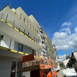 Wielopiętrowy budynek w trakcie prac elewacyjnych, widoczne rusztowania i podnośnik koszowy, balkony z żółtymi zabezpieczeniami, błękitne niebo z chmurami w tle.