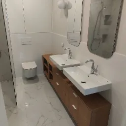 Nowoczesna łazienka z dwoma umywalkami nablatowymi na drewnianej szafce, białymi płytkami imitującymi marmur na podłodze i ścianach, oraz podwieszaną toaletą.