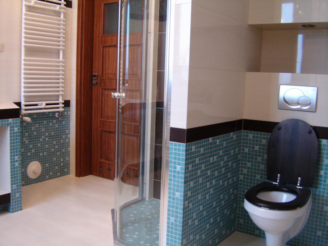 Wyremontowana łazienka z kabiną prysznicową z przezroczystego szkła, toaletą z deską w kolorze ciemnego drewna, grzejnikiem drabinkowym i mozaiką w odcieniach błękitu, wykończona listwą w kolorze...