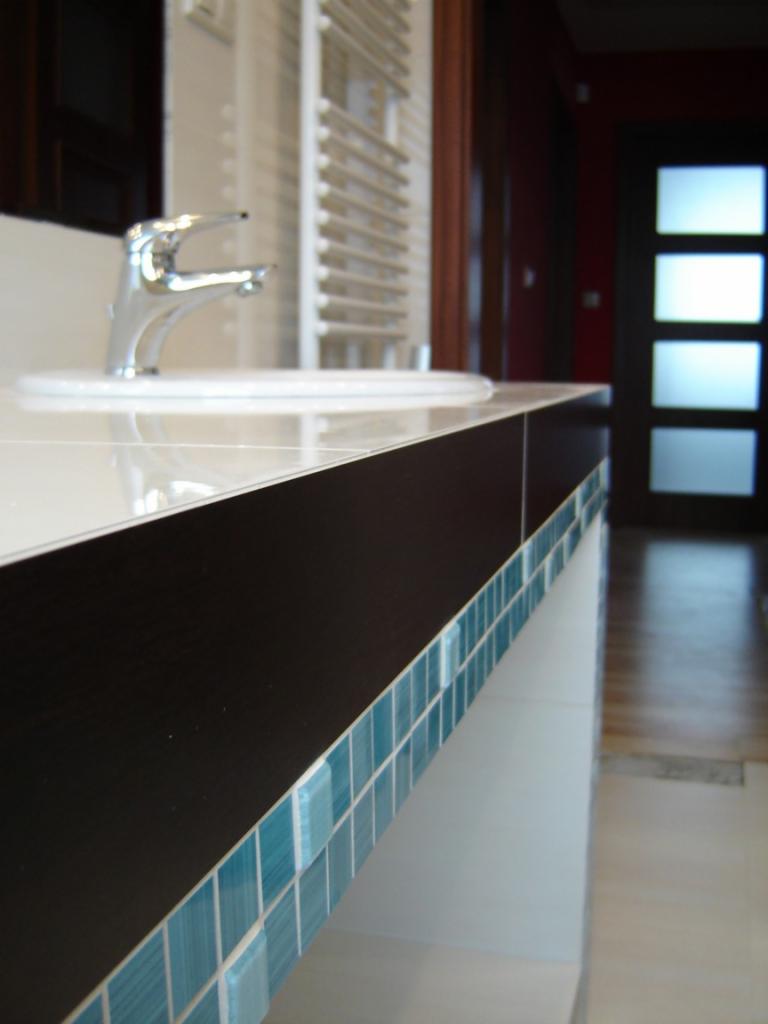 Umywalka z chromowaną baterią na blacie z ciemnego drewna, wykończonym mozaiką z błękitnych płytek; w tle grzejnik drabinkowy i drzwi z przeszkleniami.
