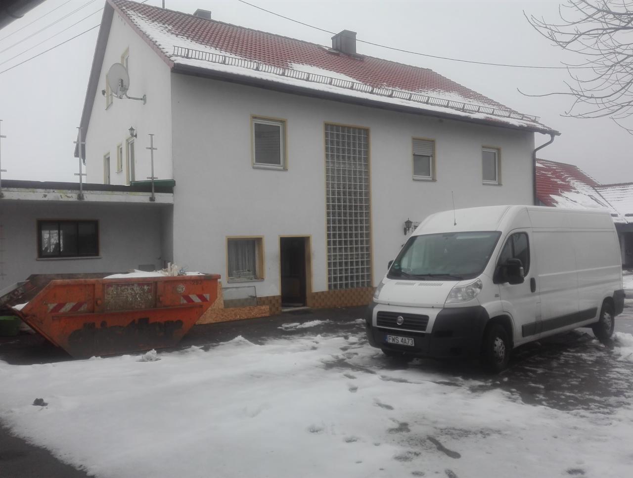 Dom jednorodzinny w trakcie remontu z pomarańczowym kontenerem na gruz i białym busem na ośnieżonym podjeździe w pochmurny dzień.