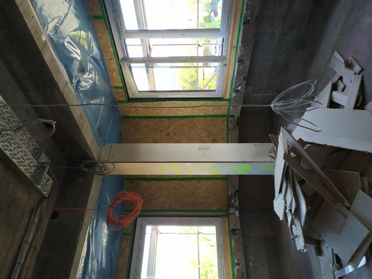 Wnętrze w trakcie rozbudowy z widocznymi oknami, izolacją, elementami konstrukcyjnymi i przewodami elektrycznymi, z fragmentami płyt kartonowo-gipsowych na podłodze.
