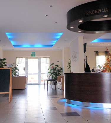 Recepcja hotelowa z drewnianą ladą podświetloną na niebiesko, roślinami doniczkowymi i oświetleniem sufitowym LED.