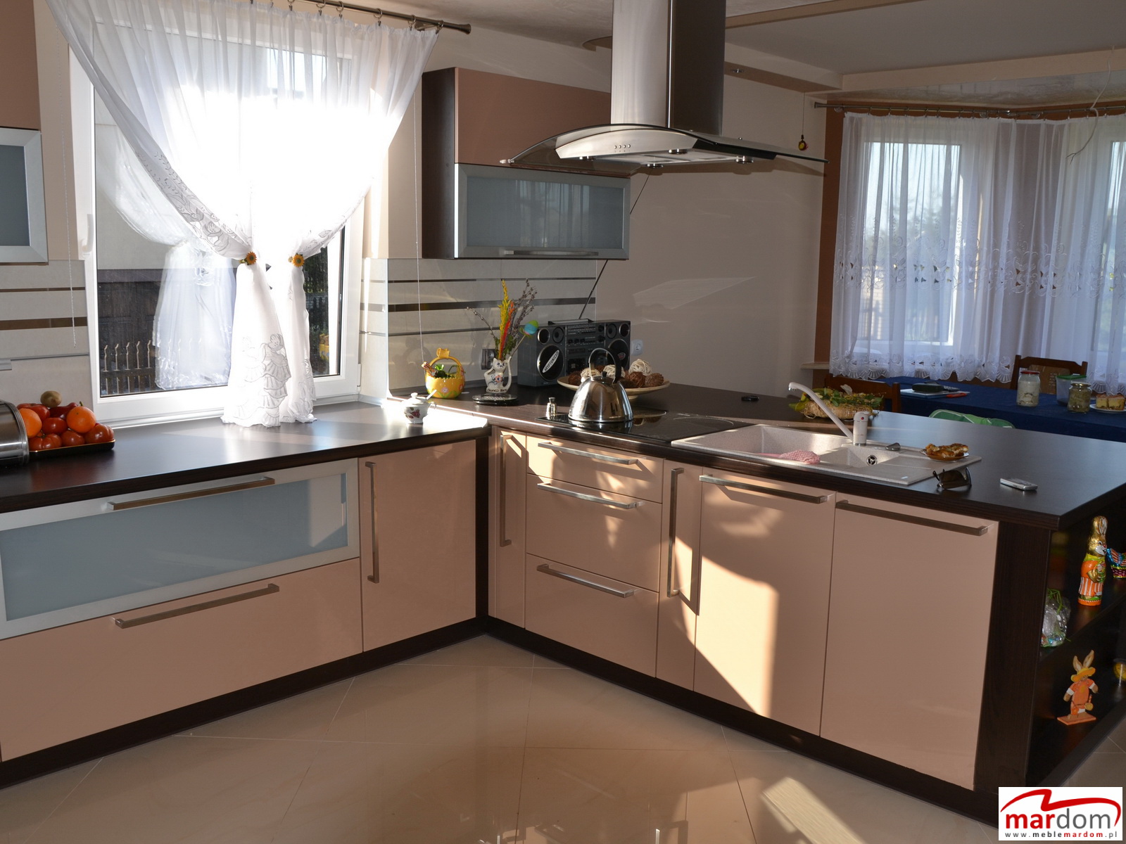 Jasna kuchnia w odcieniach beżu i brązu, z widokiem na okno z firankami i stół jadalny w tle, oświetlona popołudniowym słońcem, z czajnikiem na płycie i owocami na blacie.