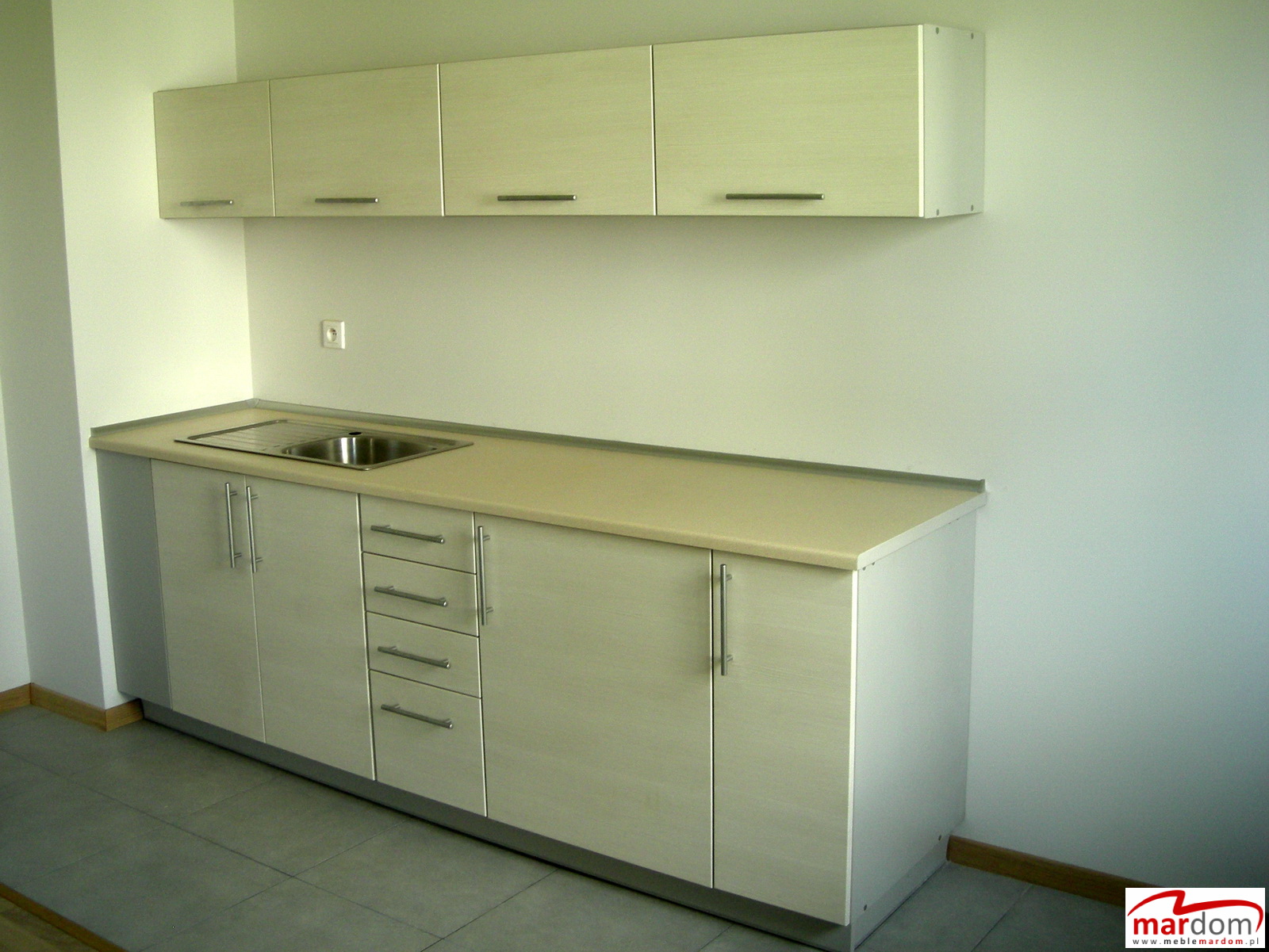 Jasna zabudowa kuchenna z górnymi szafkami i blatem ze zlewem, widok z boku na szarej podłodze.