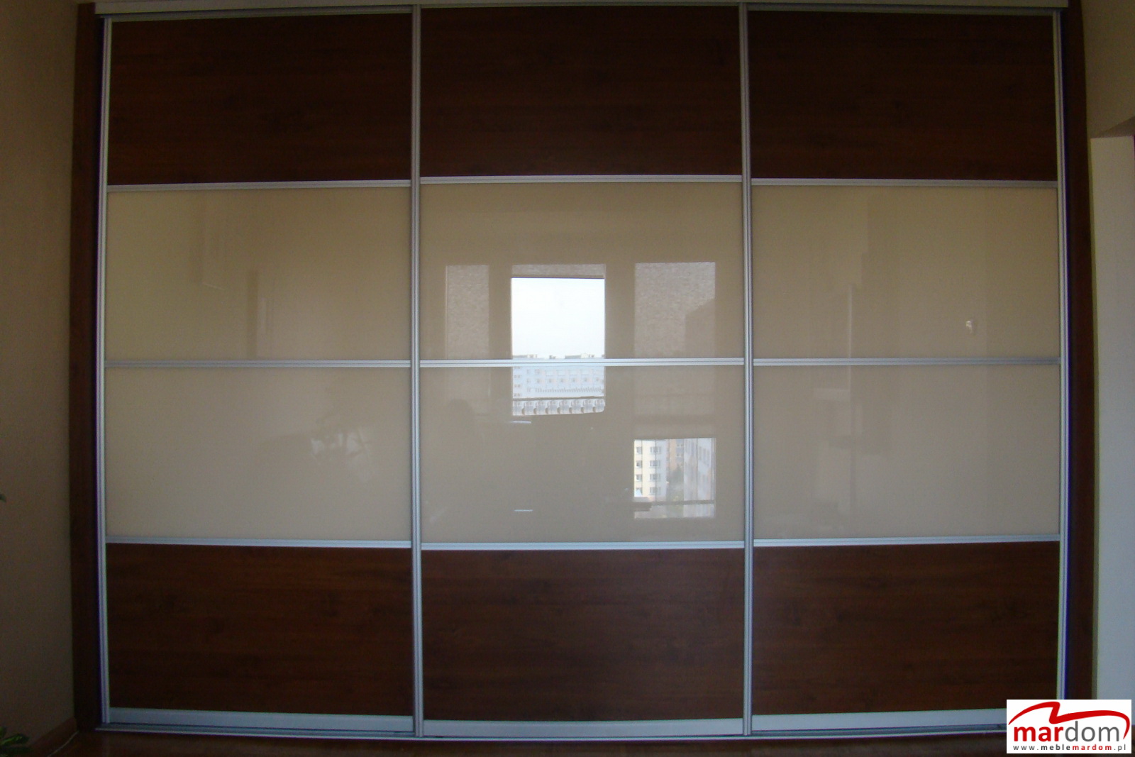 Trzydrzwiowa szafa wnękowa z przesuwnymi drzwiami, górne i dolne panele w kolorze ciemnego drewna, środkowe panele w odcieniach beżu, odbicie okna w panelach.