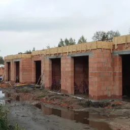 Wznoszone ściany z czerwonej cegły porotherm, widoczne otwory okienne i drzwiowe, drewniane szalunki nadproży, teren budowy po deszczu.