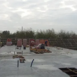 Wylewka betonowa stropu domu w trakcie budowy, widoczne zbrojenia, palety z cegłami, betoniarka i pracownicy w odblaskowych kamizelkach na tle konstrukcji więźby dachowej i drzew