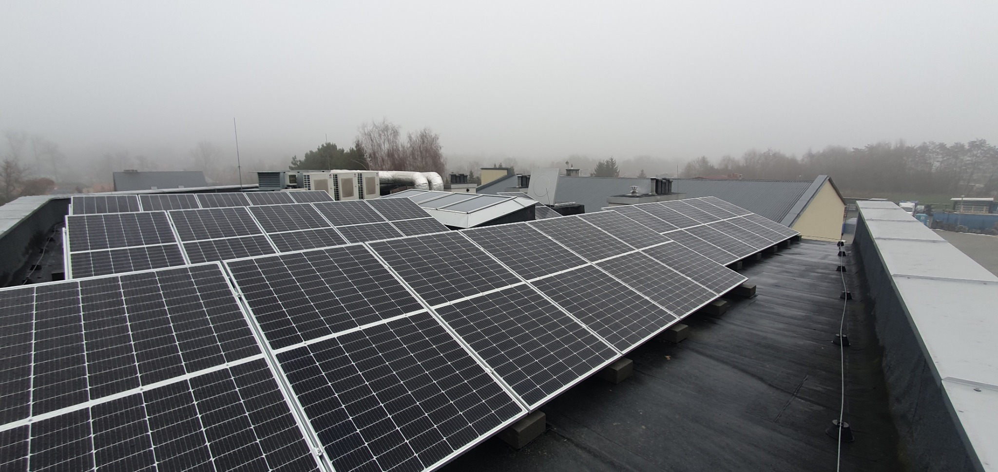 Rozległa instalacja fotowoltaiczna na płaskim dachu budynku przemysłowego w mglisty dzień, widoczne panele skierowane w stronę słońca, w tle inne budynki i drzewa.