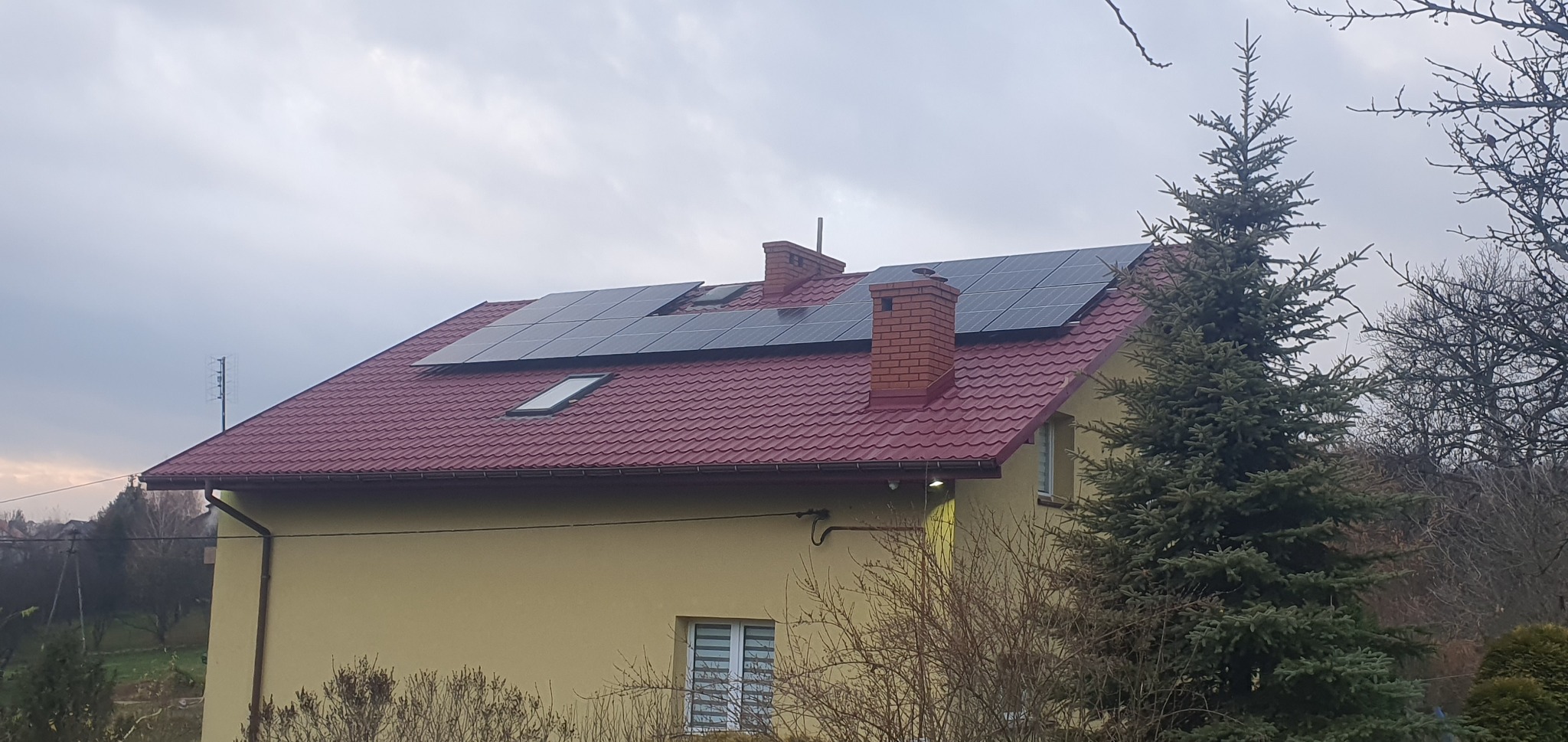 Dom jednorodzinny z panelami słonecznymi na dachu z czerwonej blachodachówki, widoczne kominy i okno dachowe, na pierwszym planie iglak i krzewy bezlistne, pochmurne niebo.