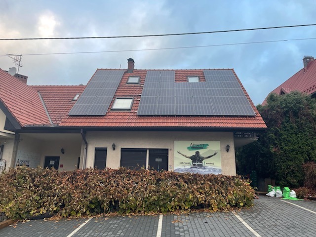 Panele słoneczne na dachu budynku z czerwoną dachówką i logo firmy na elewacji, widok z poziomu ulicy w pochmurny dzień.