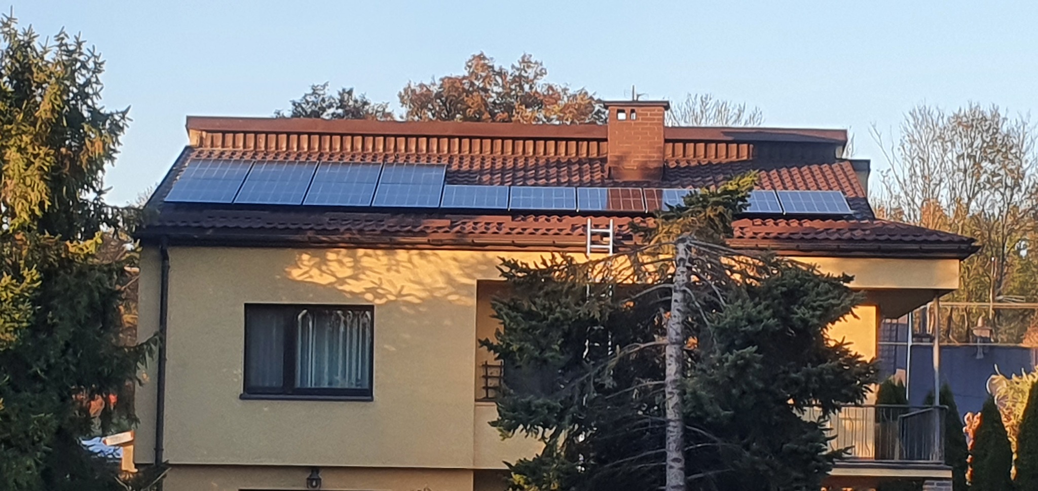 Dom jednorodzinny z brązowym dachem pokrytym panelami słonecznymi, z drabinką opartą o dach przy kominie, widoczne drzewa w otoczeniu.