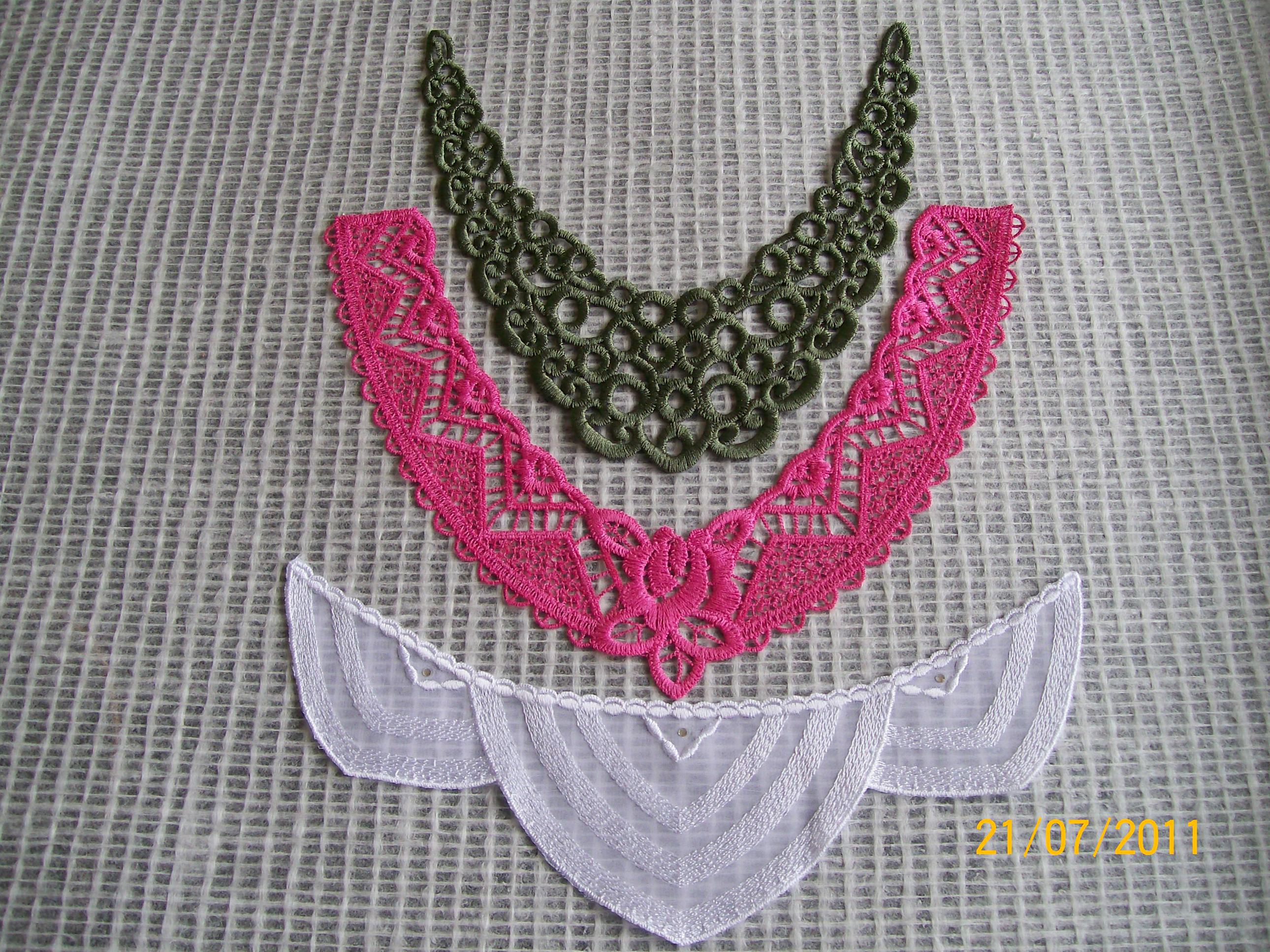 Trzy koronkowe kołnierzyki w kolorach zielonym, różowym i białym, ułożone na szarej teksturowanej tkaninie. Każdy kołnierzyk ma inny wzór i styl.