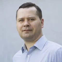 Portret mężczyzny w niebieskiej koszuli w paski, z krótkimi ciemnymi włosami i niebieskimi oczami, na jasnoszarym tle.