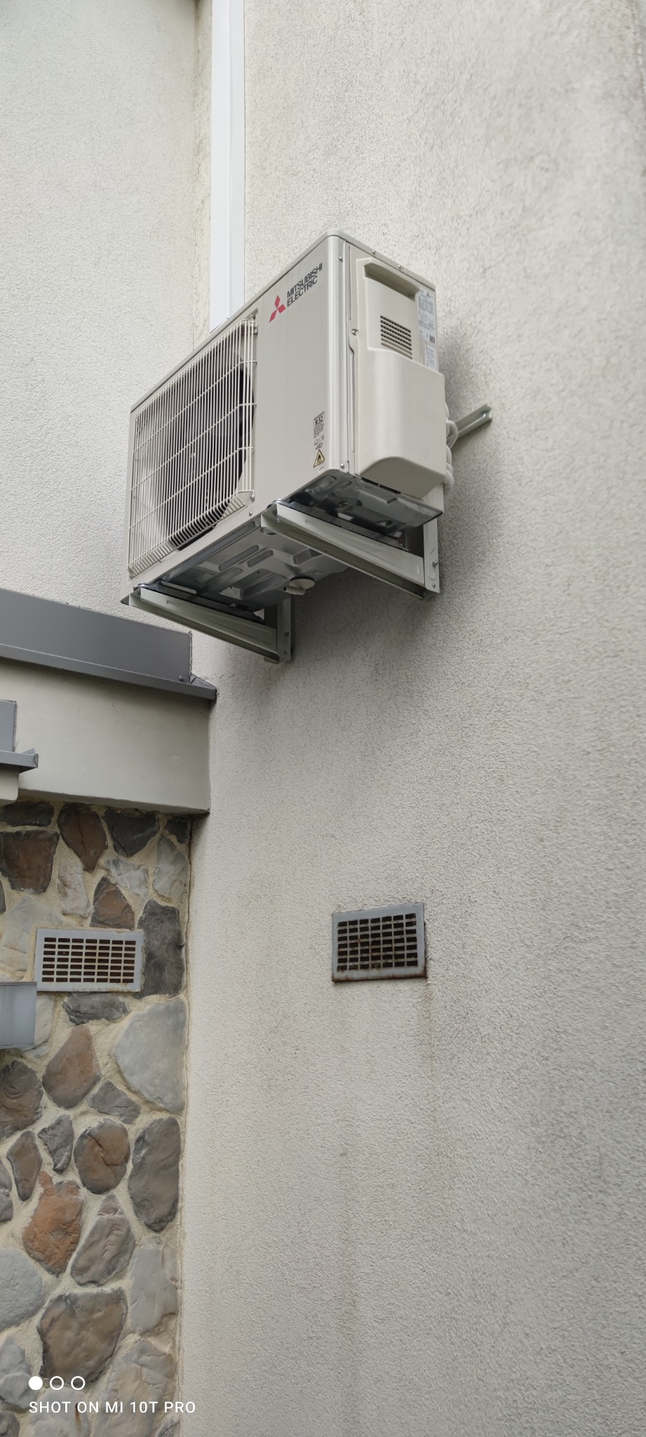 Zewnętrzna jednostka klimatyzacji Mitsubishi Electric zamontowana na wspornikach na teksturowanej, jasnej ścianie domu. Widoczne kratki wentylacyjne w ścianie i fragment elewacji z kamienia.