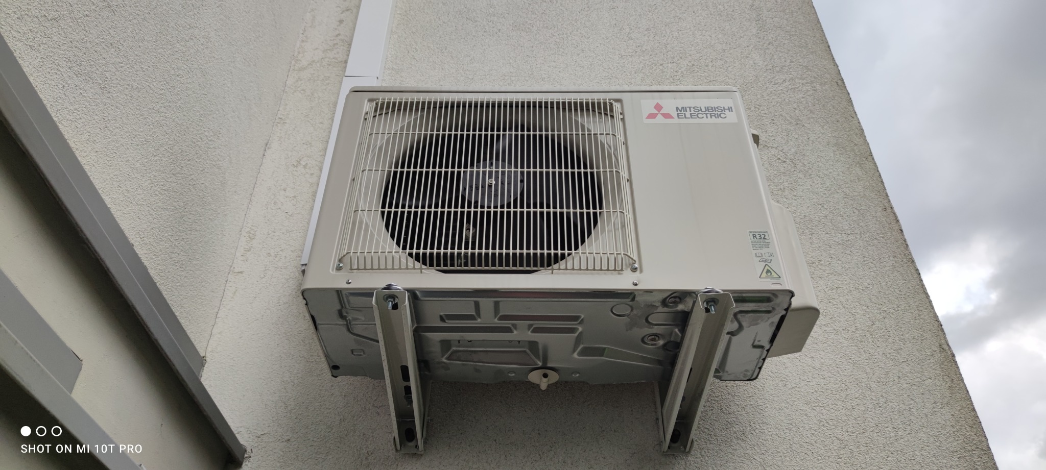 Zewnętrzna jednostka klimatyzacji Mitsubishi Electric zamontowana na ścianie budynku z widocznymi wspornikami montażowymi i etykietą R32.