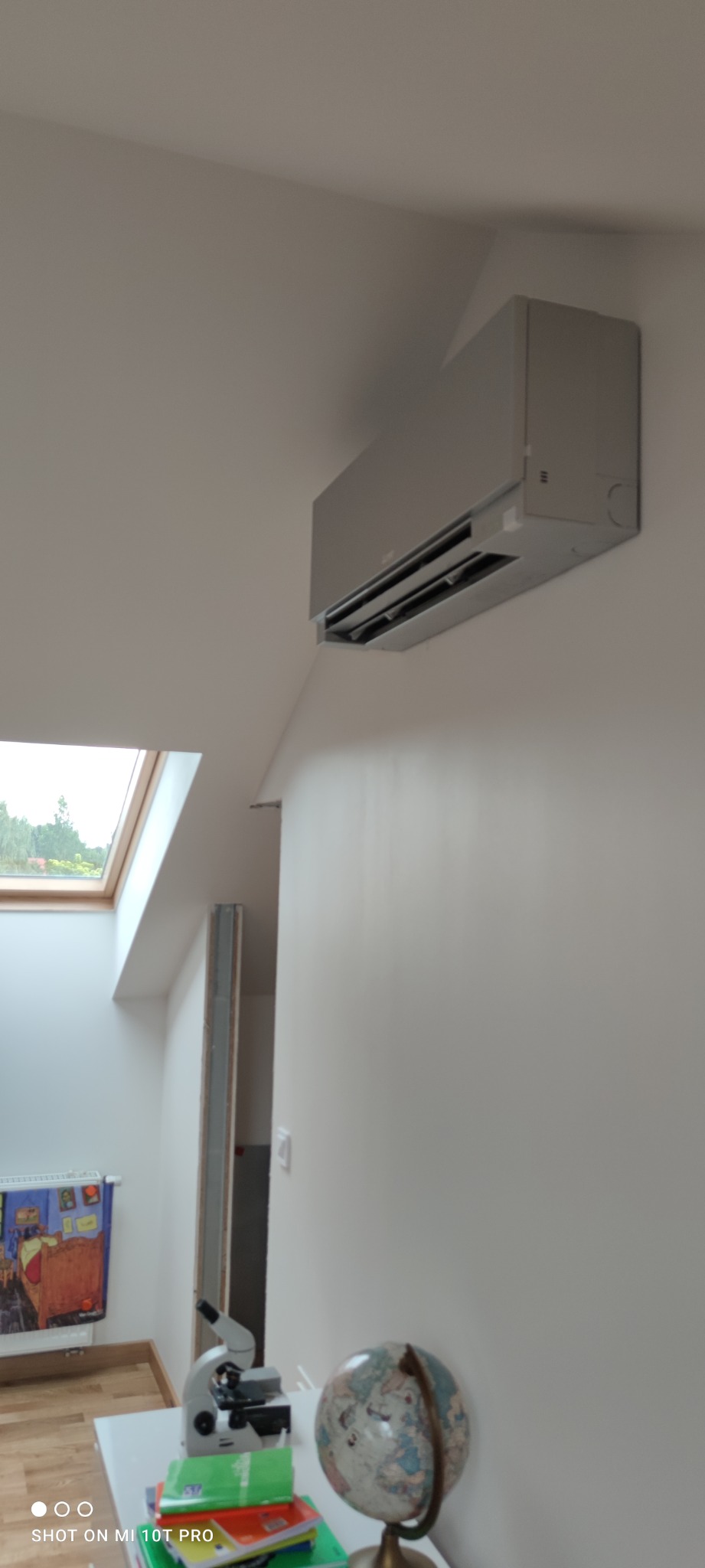 Szary klimatyzator ścienny zamontowany w pomieszczeniu z oknem dachowym i biurkiem z mikroskopem i globusem.