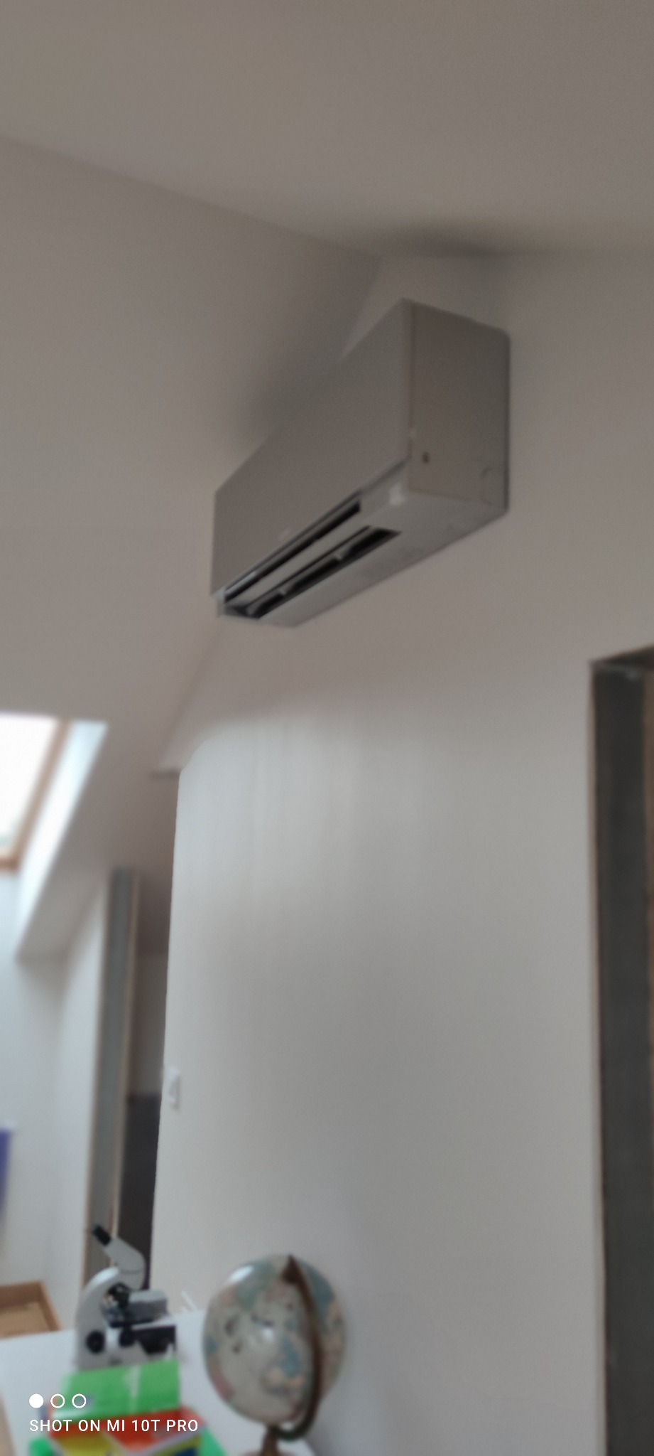 Szary klimatyzator ścienny zamontowany w pokoju z oknem dachowym, widoczny mikroskop i globus na biurku w tle.
