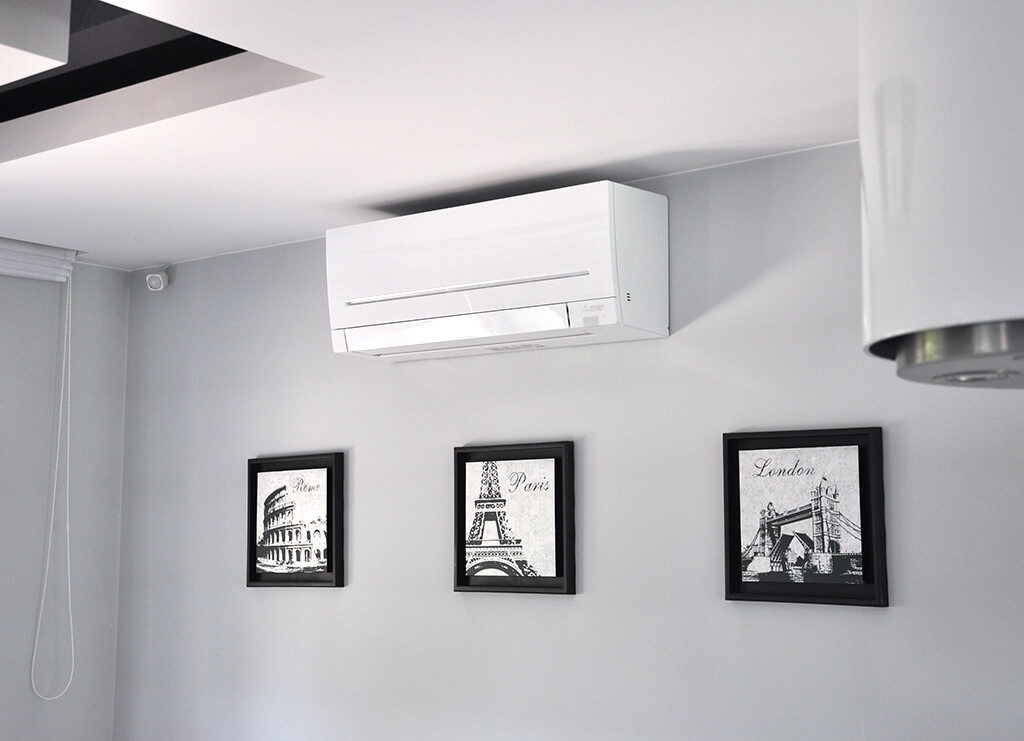 Biały klimatyzator ścienny Mitsubishi Electric w nowoczesnym wnętrzu z szarymi ścianami, trzema obrazami w czarnych ramkach przedstawiającymi Koloseum, Wieżę Eiffla i Tower Bridge.