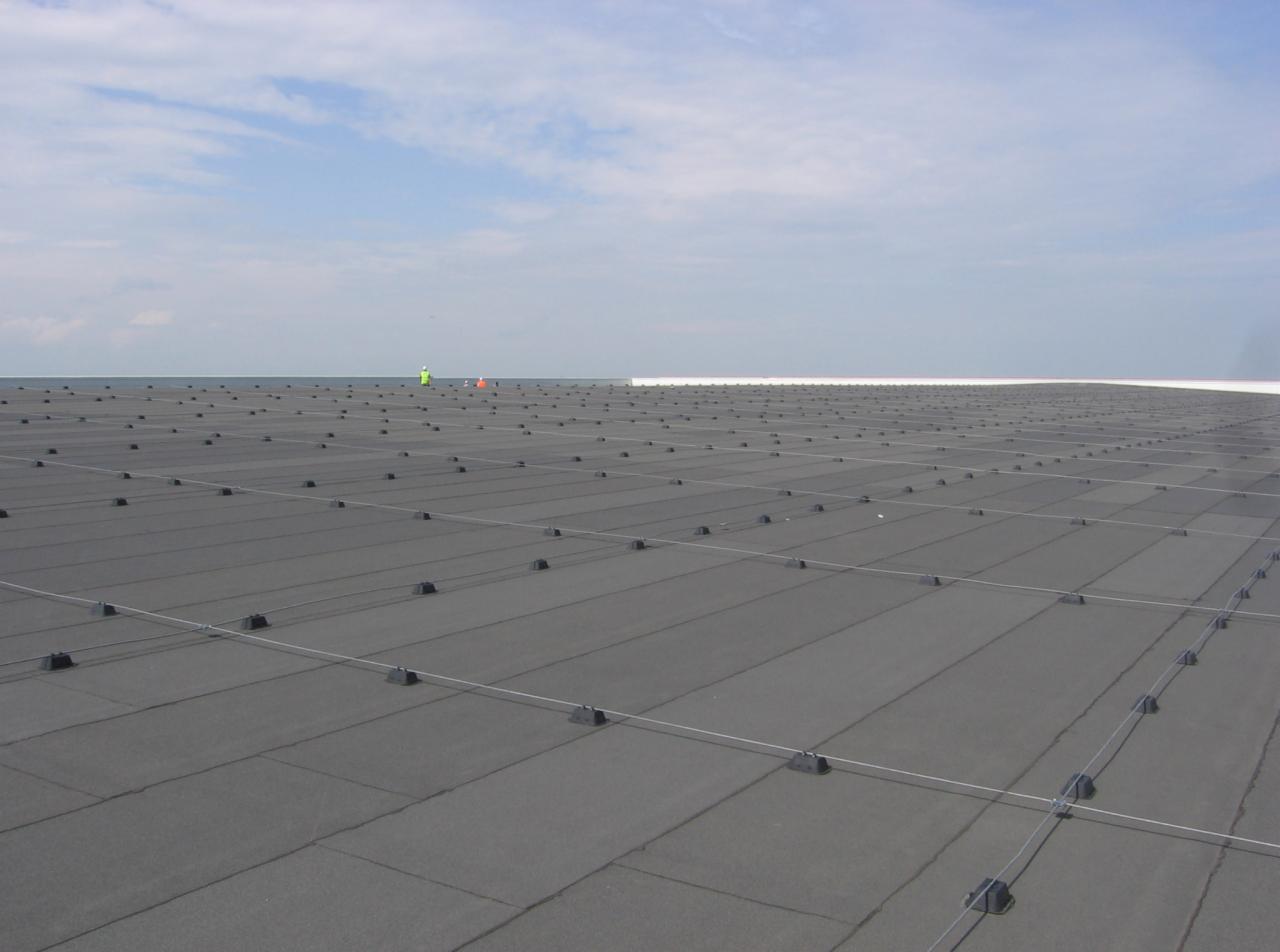 Rozległy, płaski dach pokryty ciemną membraną z regularnym rozmieszczeniem elementów mocujących i linkami zabezpieczającymi, w tle widoczni pracownicy w odblaskowych ubraniach.