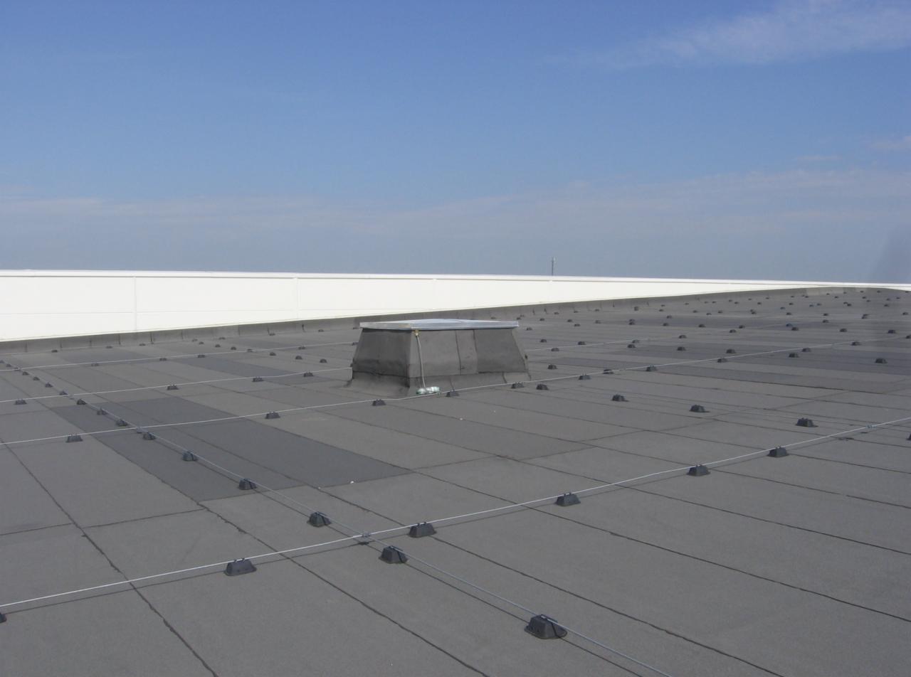 Płaski dach pokryty szarą membraną z widocznym świetlikiem i systemem odgromowym, na tle błękitnego nieba.
