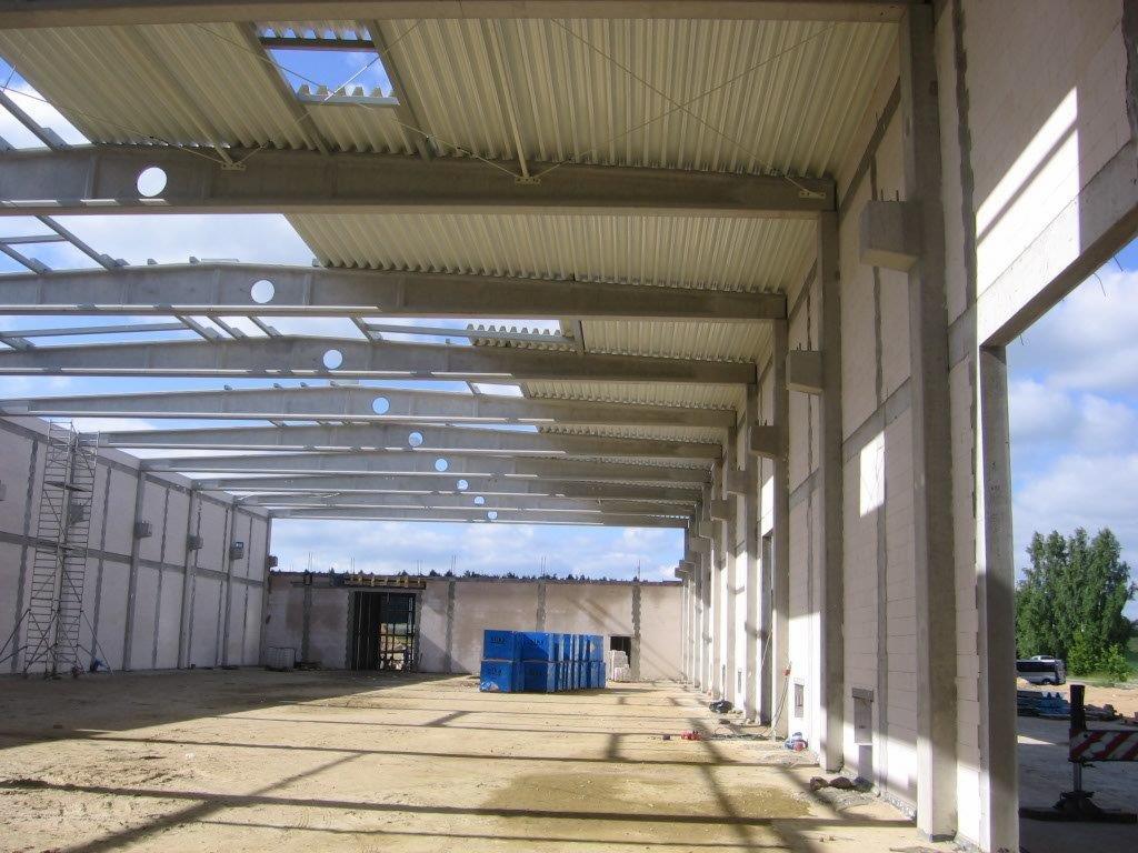 Wnętrze hali w trakcie budowy z widoczną konstrukcją stalową dachu i ścianami z betonu komórkowego, niekompletnym poszyciem dachowym z blachy falistej z widocznym niebem przez otwory...