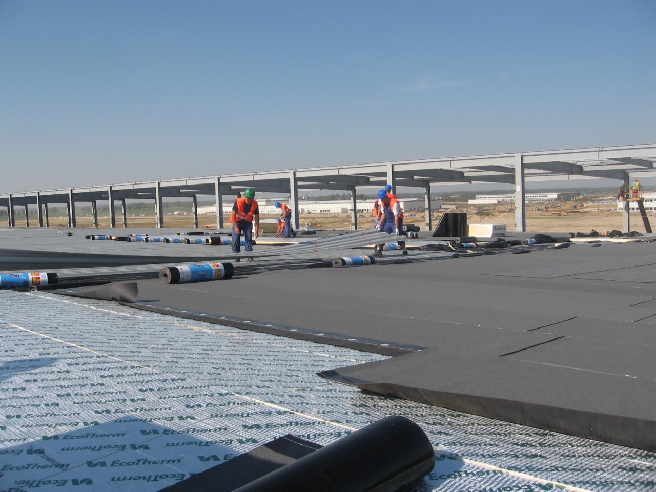 Prace na dachu hali przemysłowej: układanie izolacji termicznej EcoTherm przez robotników w kaskach i kamizelkach odblaskowych, z widoczną konstrukcją stalową dachu i odległym placem budowy w tle.