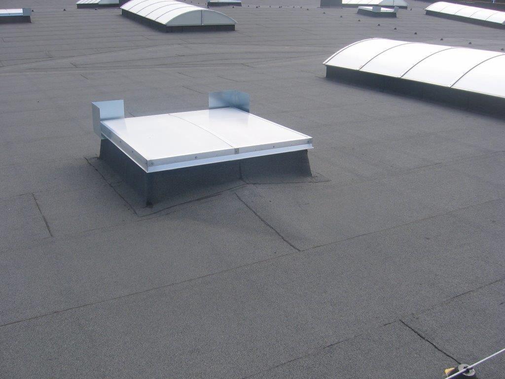 Płaski dach pokryty papą z zamontowanymi świetlikami dachowymi o różnych kształtach, w tym kwadratowym z metalowymi obudowami.